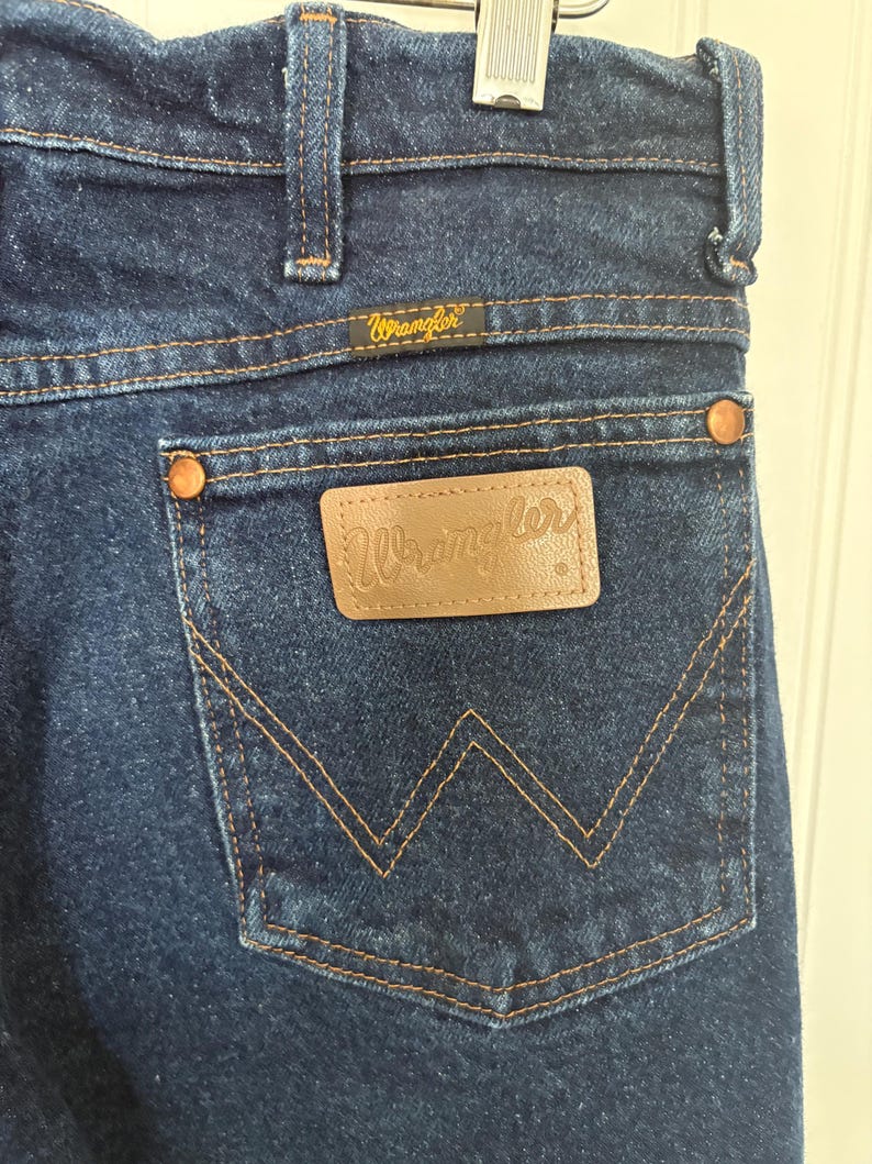 May include: Close-up of dark blue denim jeans with the Wrangler logo on the back pocket and waistband. The jeans have copper-colored rivets and a classic five-pocket design. The stitching is a contrasting tan color.