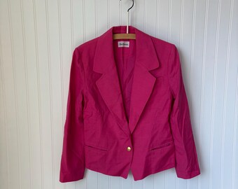 May include: A bright pink blazer with a single gold button closure. The blazer has a classic notched lapel and two front pockets. It is hanging on a wooden hanger against a white paneled wall.