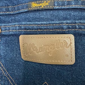 May include: Close-up of the back pocket of blue denim jeans. A brown leather patch with the embossed word "Wrangler" is visible. Above the pocket is a small tag with the word "Wrangler" in gold lettering.