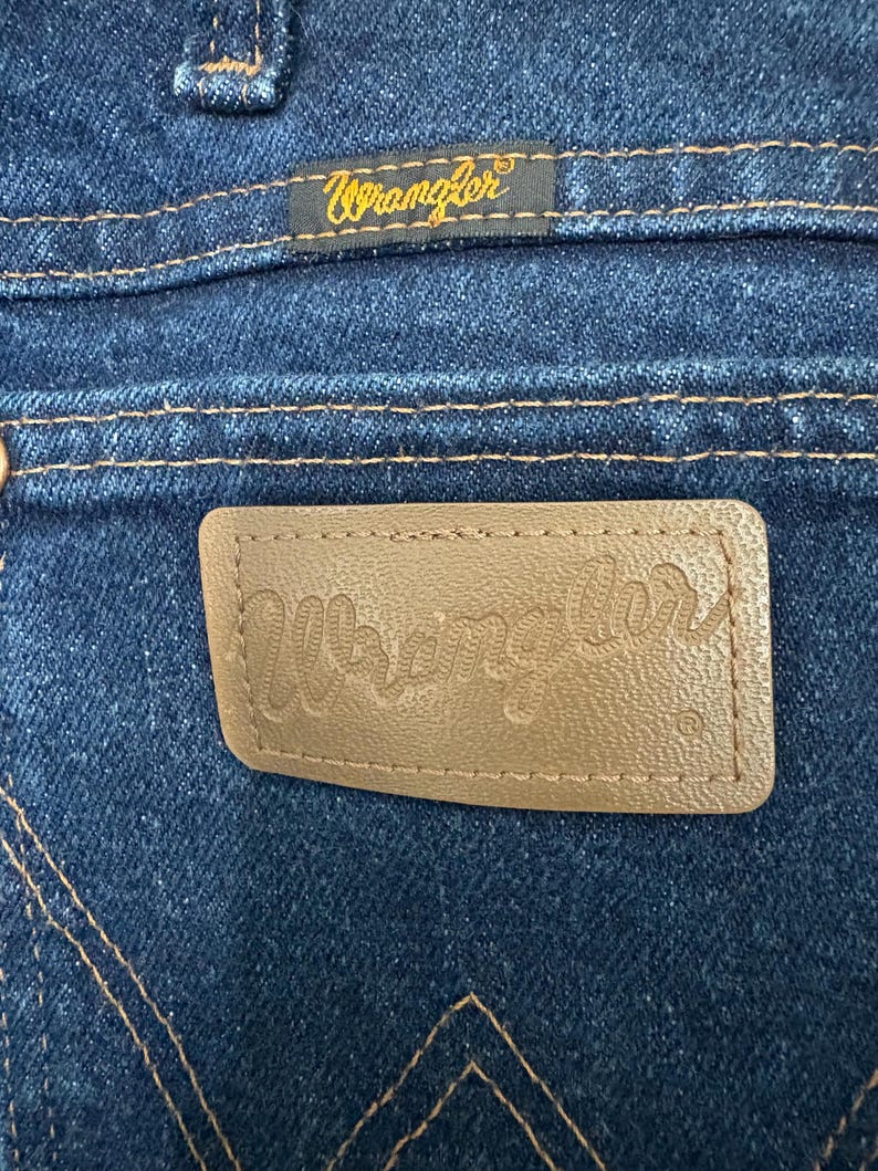 May include: Close-up of the back pocket of blue denim jeans. A brown leather patch with the embossed word "Wrangler" is visible. Above the pocket is a small tag with the word "Wrangler" in gold lettering.