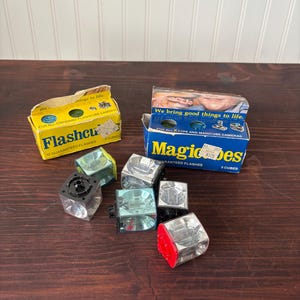 May include: Vintage flashcubes and boxes. The boxes are yellow and blue with text that reads "Flashcube" and "Magicubes". The flashcubes are clear with black and red accents. The boxes and flashcubes are on a dark wooden surface.