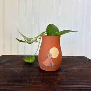 May include: A terracotta vase featuring a hand-painted teepee and sun design. The vase contains a green plant with large leaves. The vase sits on a dark wooden surface, set against a white backdrop.