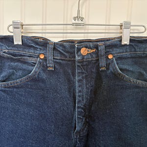 May include: Close-up of a pair of dark blue denim jeans hanging on a metal hanger. The jeans have copper-colored rivets and a button closure. The pockets and seams are visible, showcasing the classic design.