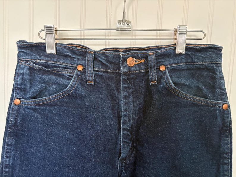 May include: Close-up of a pair of dark blue denim jeans hanging on a metal hanger. The jeans have copper-colored rivets and a button closure. The pockets and seams are visible, showcasing the classic design.