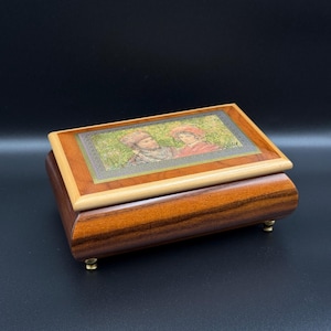 May include: A rectangular wooden jewelry box with a hinged lid. The lid features a framed image of two people in period clothing. The box is made of dark brown wood with a lighter wood trim and rests on small brass feet.