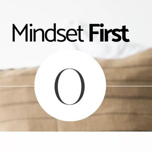 May include: A white circle with the letter "O" in black text. The text "Mindset First" is above the circle in black text. The background is a beige pillow with a white surface behind it.