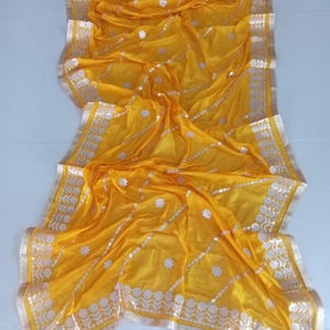 May include: A yellow sheer saree with a silver and gold embroidered border. The saree has a floral pattern and is perfect for special occasions.