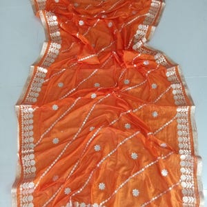 May include: Orange sheer fabric saree with silver and white embroidery. The saree has a floral design and a border with a paisley pattern.