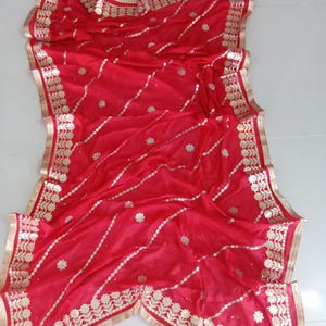 May include: Red saree with gold embroidery and a decorative border. The saree is made of a sheer fabric and has a delicate floral pattern.