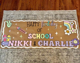 Puede incluir: Una pancarta marrón con las palabras "HAPPY first day OF SCHOOL NIKKI & CHARLIE" en letras de burbujas coloridas. La pancarta presenta un signo de paz, un lápiz, crayones, un arcoíris, flores y estrellas.