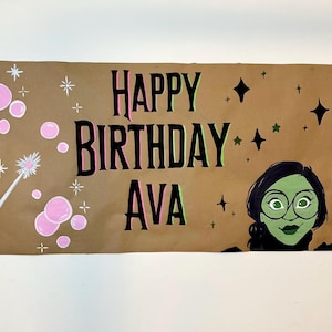 Może przedstawiać: Brązowy papierowy baner z napisem "Happy Birthday Ava" napisanym czarnym kolorem z różowymi i zielonymi obramowaniami. Baner zawiera również kreskówkową ilustrację dziewczyny o zielonej skórze, czarnych włosach i okularach. Na banerze znajdują się białe gwiazdki i różowe bąbelki.