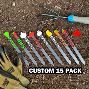 May include: A set of 10 garden plant markers with different vegetable illustrations and names. The markers are made of grey plastic and have a pointed end for easy insertion into the soil. The markers are arranged in a row on a bed of brown soil. The text "CUSTOM 15 PACK" is printed in white on the soil.