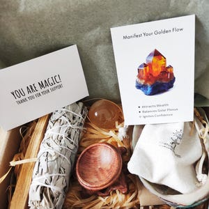 Peut inclure: Coffret cadeau contenant un fagot de sauge blanche, un bol en bois, un cristal, une petite pochette et une carte avec l'inscription "Manifestez votre flux doré". La boîte contient également une carte avec l'inscription "Vous êtes magique !"