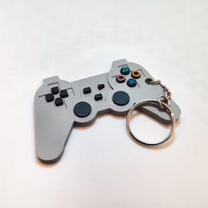 May include: A gray video game controller keychain with black buttons and colorful action buttons. The keychain has a silver key ring attached. The controller design is a classic style.