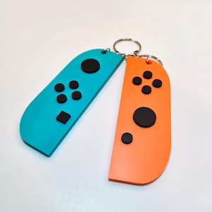 May include: Two keychains shaped like Nintendo Switch Joy-Con controllers. One is teal, the other is orange. Each has black buttons and a silver keyring. The controllers are a fun accessory for gamers.