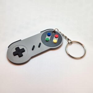 May include: A gray keychain shaped like a classic video game controller. The controller has a black directional pad, colored buttons, and a silver chain with a keyring. The design is a nostalgic nod to retro gaming.