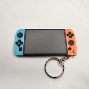 May include: A keychain shaped like a video game console. The keychain has a black screen, with blue and orange sides. It features black buttons and a silver keyring.