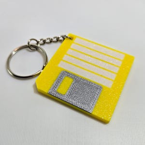 May include: A yellow floppy disk keychain with a silver metal slider and a silver metal ring. The keychain is made of a textured material.