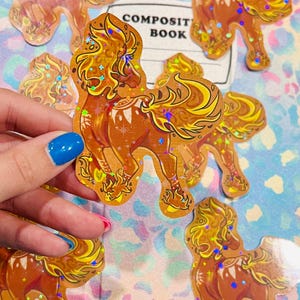 May include: A collection of holographic stickers featuring a stylized horse design with a flowing mane and tail. The stickers are primarily brown and gold, with iridescent accents. The word "COMPOSIT BOOK" is visible in the background.