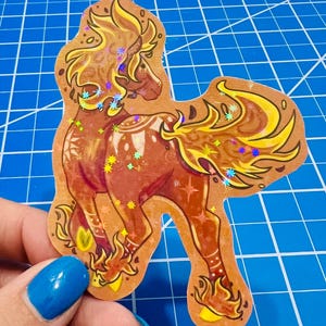 May include: A brown sticker featuring a stylized horse with a flowing mane and tail in shades of yellow and gold. The horse has a dark red body and is decorated with holographic star-shaped accents. The sticker is cut in the shape of the horse.