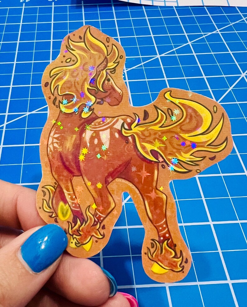 May include: A brown sticker featuring a stylized horse with a flowing mane and tail in shades of yellow and gold. The horse has a dark red body and is decorated with holographic star-shaped accents. The sticker is cut in the shape of the horse.
