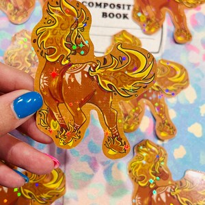 May include: Holographic stickers featuring a stylized horse with a flowing mane and tail. The stickers are primarily brown and gold with iridescent accents. The text "COMPOSIT BOOK" is visible in the background. These stickers are ideal for scrapbooking and crafts.