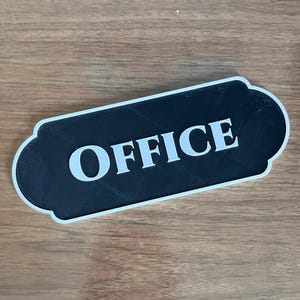 Puede incluir: Un letrero de oficina en blanco y negro con un borde decorativo. La palabra "OFFICE" está impresa en letras blancas en negrita sobre un fondo negro. El letrero es rectangular con esquinas redondeadas.