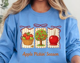 May include: A light blue sweatshirt featuring a graphic with three panels: a basket of apples, an apple tree, and a caramel apple. The text "Apple Pickin' Season" is below the panels. The sweatshirt is worn by a person.