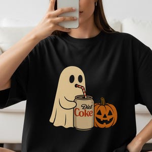 Puede incluir: Camiseta negra con un fantasma de dibujos animados bebiendo de una lata de Diet Coke con una pajita, junto a una calabaza naranja tallada. El fantasma tiene dos ojos negros y sostiene la lata. Las palabras "Diet Coke" están impresas en la lata.