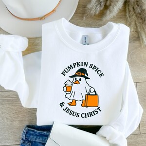 May include: White sweatshirt with a graphic of a ghost wearing a witch hat, holding a pumpkin spice latte and a shopping bag. The text reads "Pumpkin Spice & Jesus Christ". The sweatshirt is folded on a wooden surface.