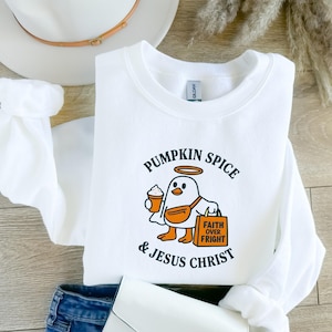 May include: White sweatshirt with a graphic of a cartoon bird holding a pumpkin spice latte and a bag that says "Faith Over Fright." The text "Pumpkin Spice & Jesus Christ" is arched above and below the bird. A white hat and purse are in the background.