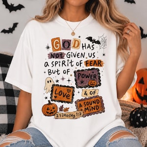 May include: A white short-sleeve t-shirt featuring a graphic print with the text "GOD HAS Not Given Us a SPIRIT of FEAR But of POWER & OF A SOUND MIND 2 Timothy 1:7". The design includes illustrations of a ghost, pumpkins, bats, and a spiderweb.