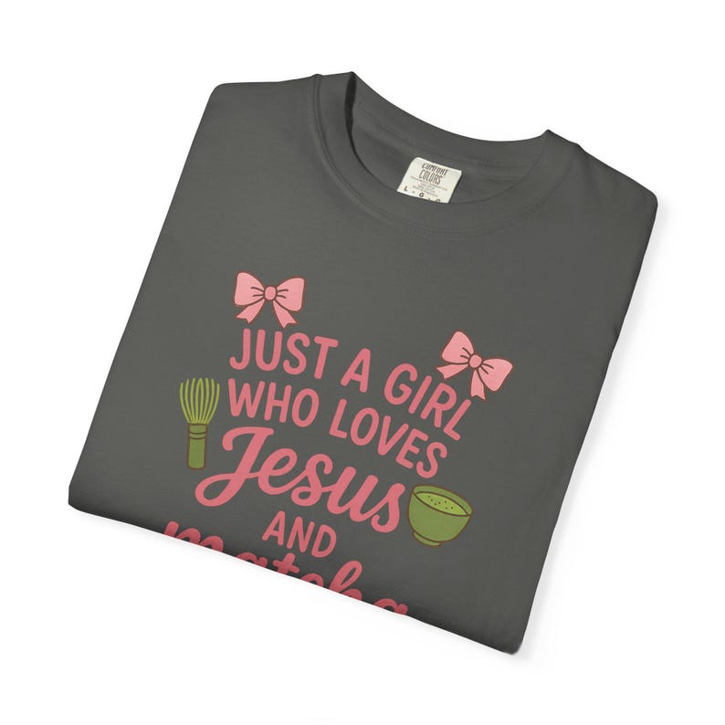 May include: Dark gray t-shirt with the text "JUST A GIRL WHO LOVES Jesus AND matcha" in pink. The design includes pink bows, a matcha whisk, and a matcha bowl.
