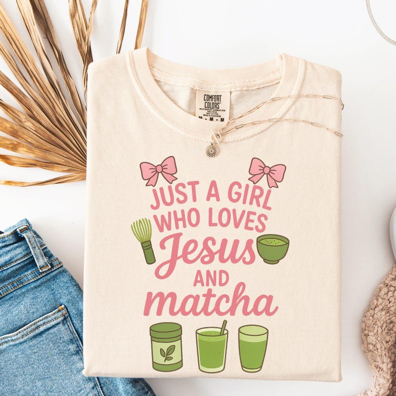 May include: Cream-colored t-shirt with the text "JUST A GIRL WHO LOVES Jesus AND matcha" in pink and green. The shirt features pink bows, a matcha whisk, and matcha illustrations. A gold necklace and blue jeans are visible.
