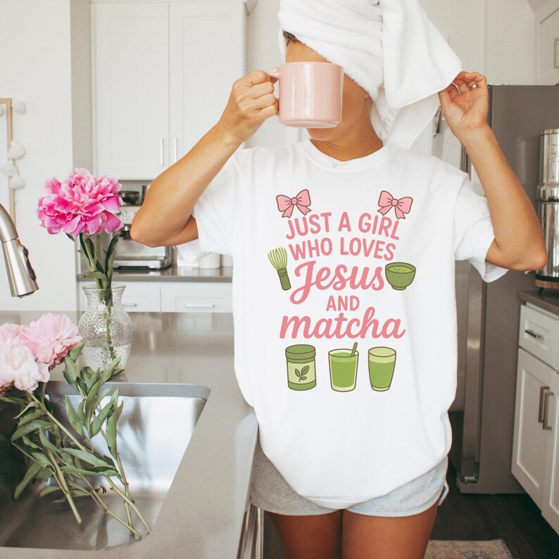 May include: White t-shirt with the text "Just a Girl Who Loves Jesus and Matcha" in pink and green. The shirt features illustrations of a whisk, a bowl, and matcha drinks. A person is holding a pink mug.