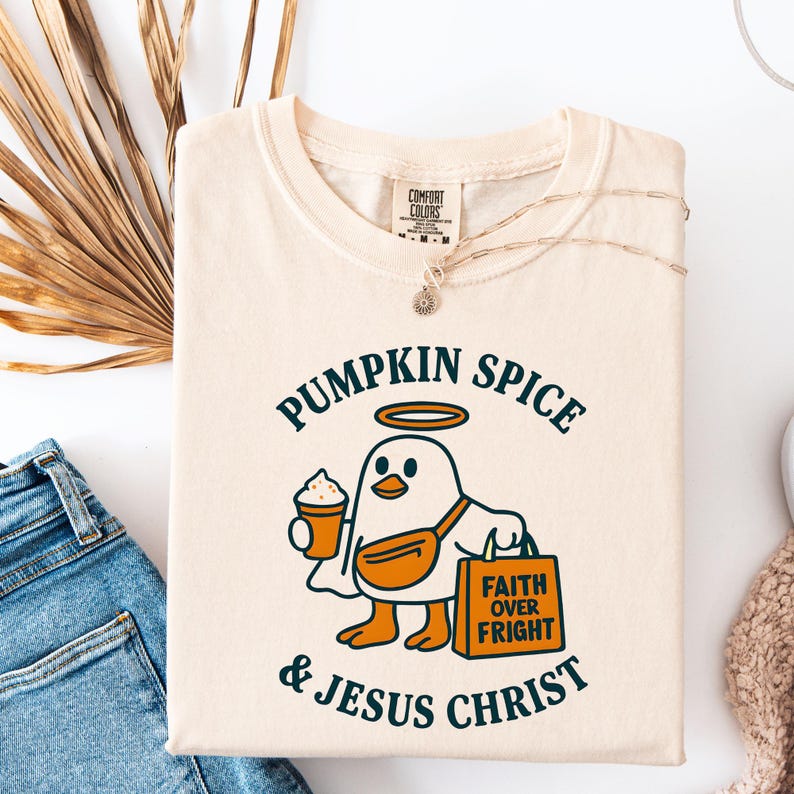 May include: Cream-colored t-shirt with a graphic of a cartoon bird holding a coffee cup and a shopping bag. The text on the shirt reads "PUMPKIN SPICE & JESUS CHRIST" and "FAITH OVER FRIGHT". A gold necklace is draped on the shirt.