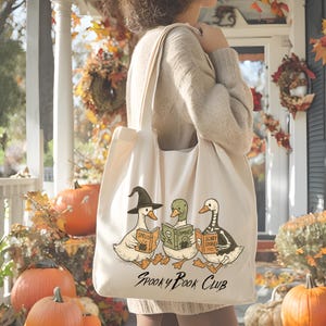 May include: Cream tote bag with a Halloween design. It shows three cartoon ducks reading books labelled "Spells", "Zombies", and "Scary Stories". The text "Spooky Book Club" is printed below. Pumpkins and autumn leaves are in the background, creating a seasonal atmosphere.