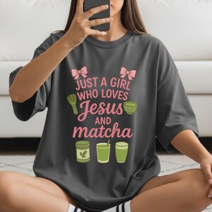 May include: Dark gray t-shirt with the text "JUST A GIRL WHO LOVES Jesus AND matcha" in pink and white. The design includes pink bows, a whisk, a bowl, and matcha-related graphics. A casual, comfortable tee.