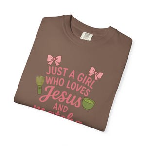 May include: A brown t-shirt with the text "JUST A GIRL WHO LOVES Jesus AND matcha" in pink. The design includes pink bows, a green matcha whisk, and a green matcha bowl.