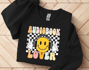 May include: Black sweatshirt with the words "AUDIOBOOK LOVER" in colorful retro font. A smiling yellow face wearing headphones is centered on a black and white checkered background, with peace signs on either side.
