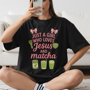 May include: Black t-shirt with the text "JUST A GIRL WHO LOVES Jesus AND matcha" in pink and white. The design includes pink bows, a matcha whisk, a bowl of matcha, and matcha drinks.