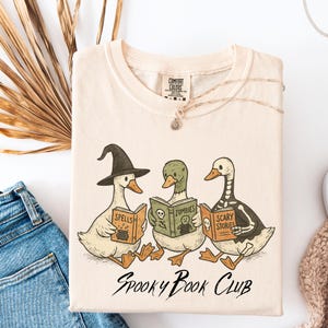 May include: Cream-colored t-shirt featuring a whimsical illustration of three ducks reading books labeled "Spells," "Zombies," and "Scary Stories." The text "Spooky Book Club" is below the illustration. A silver necklace is draped on the shirt.
