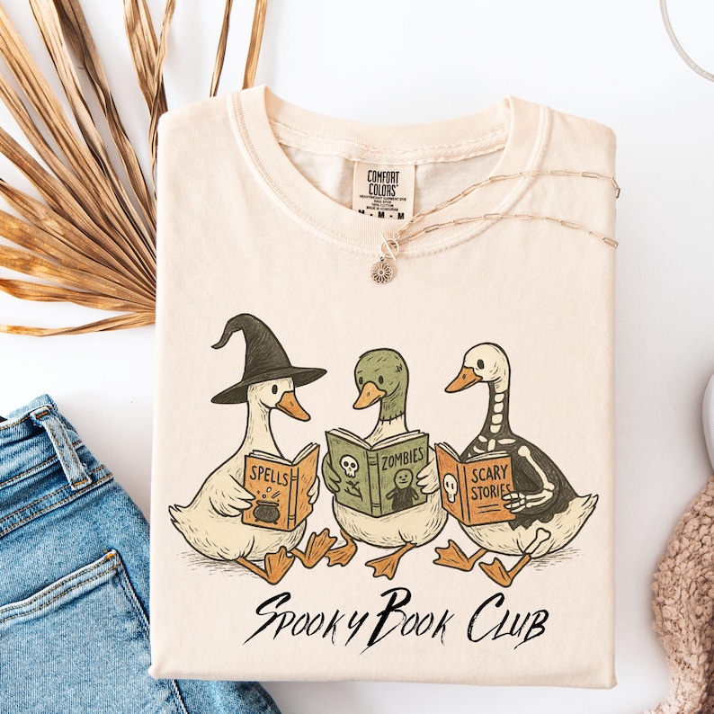May include: A cream-coloured t-shirt with a cartoon illustration of three ducks reading books labelled "Spells", "Zombies", and "Scary Stories". The text "Spooky Book Club" is below the illustration. A silver necklace is draped on the shirt.