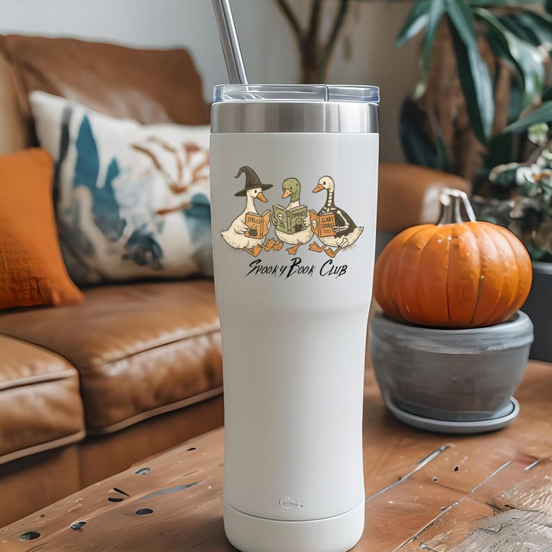 May include: A white insulated travel mug with a silver straw. The mug features a cartoon illustration of three ducks reading books, each in a Halloween costume. The text "Spooky Book Club" is below the ducks.