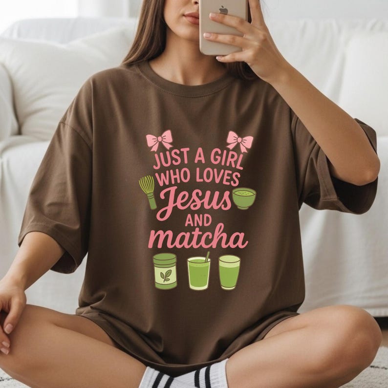 May include: A brown t-shirt with the text "JUST A GIRL WHO LOVES Jesus and matcha" in pink and green. The design includes a matcha whisk, a bowl of matcha, and matcha drinks. The shirt is a casual, comfortable style.