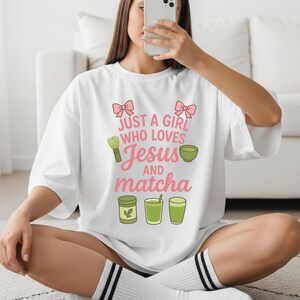 May include: White t-shirt with the text "JUST A GIRL WHO LOVES Jesus AND matcha" in pink and green. The design includes pink bows, a matcha whisk, a bowl of matcha, and matcha drinks. The shirt is a casual, oversized style.