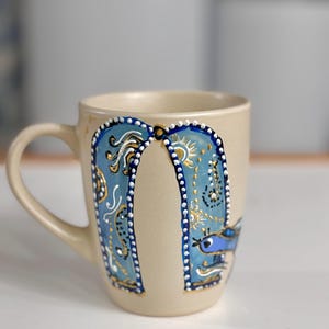 May include: A beige ceramic mug with a curved handle. The mug features a blue and gold painted design with a bird and abstract shapes. The design is framed by a border of white dots. The mug is suitable for hot or cold beverages.