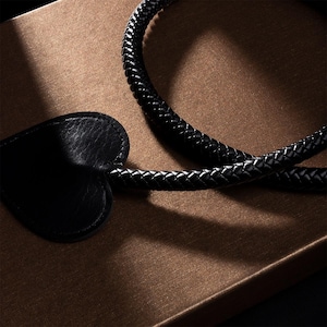 Puede incluir: Un charm de cuero negro en forma de corazón unido a un cordón de cuero trenzado negro.