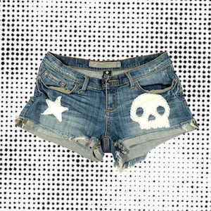 May include: Blue denim shorts with a distressed look. The shorts feature a white star and a white skull design. The edges are frayed, and the shorts have classic five-pocket styling.