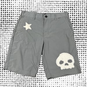 May include: Grey shorts with a button fastening and pockets. The shorts have a white star patch on the left and a white skull patch on the right. They are made from a lightweight material.